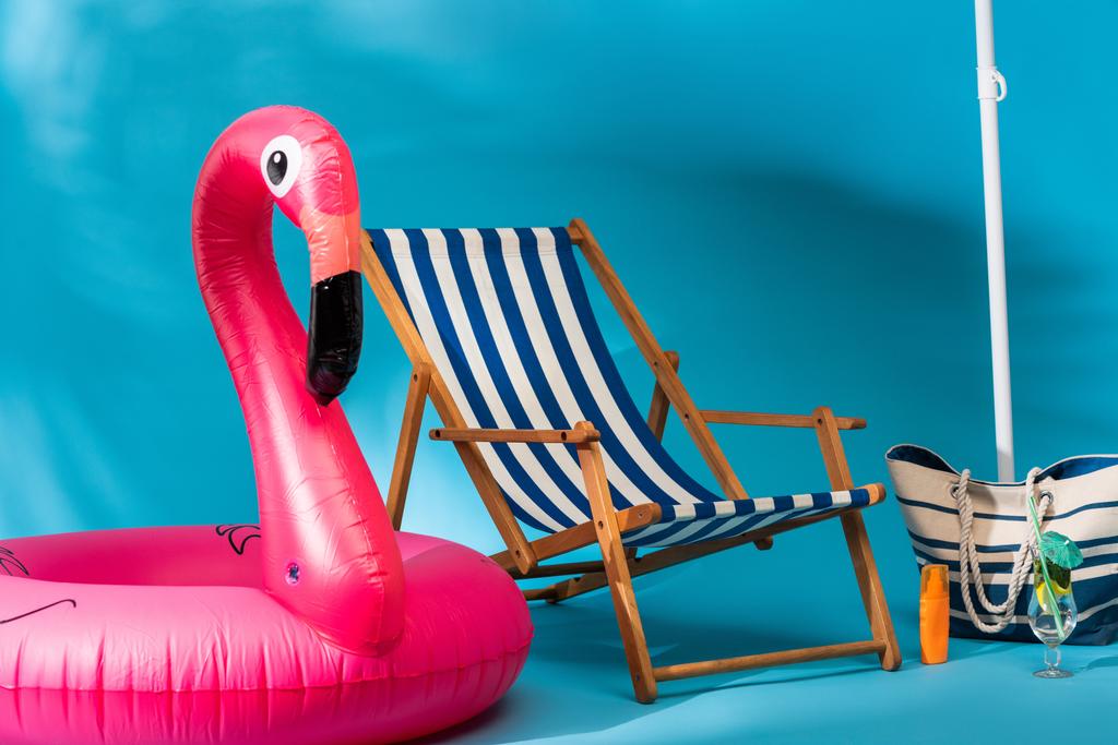 полосатый шезлонг возле надувного фламинго, солнцезащитный крем, пляжная сумка и коктейль на синем фоне - Фото, изображение