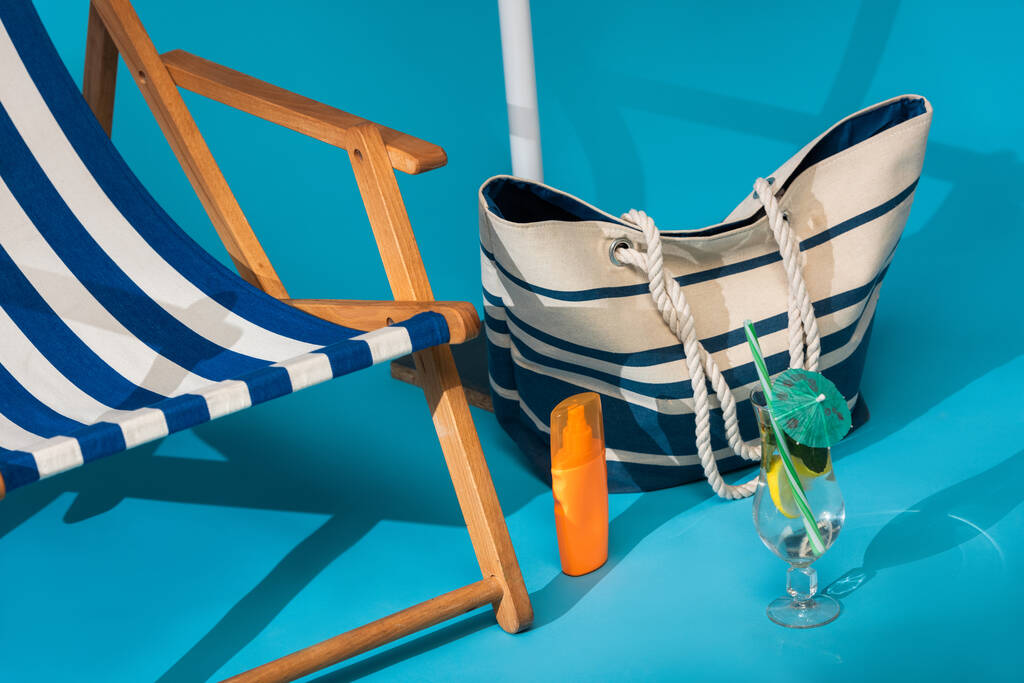 close up view of striped deck chair near sunscreen, beach bag and cocktail on blue background - Photo, Image