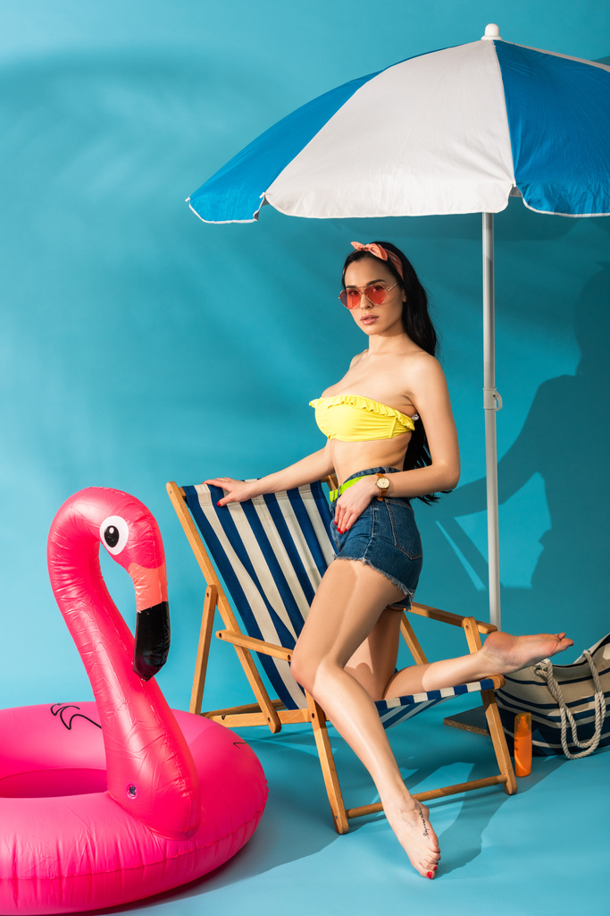 chica elegante sentada posando en la silla de cubierta cerca de flamenco inflable, bolso de playa y paraguas sobre fondo azul
 - Foto, imagen