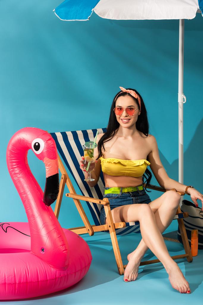 Gülümseyen stil sahibi kız güverte koltuğunda şemsiyenin altında mavi arka planda şişme flamingo altında kokteyl ile oturuyor. - Fotoğraf, Görsel