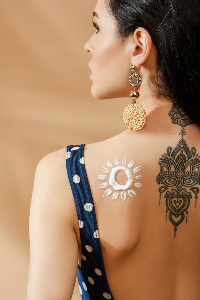 back view of sexy brunette woman in polka dot swimsuit with drawn sun made of sunscreen and tattoo on back on beige background - Photo, Image