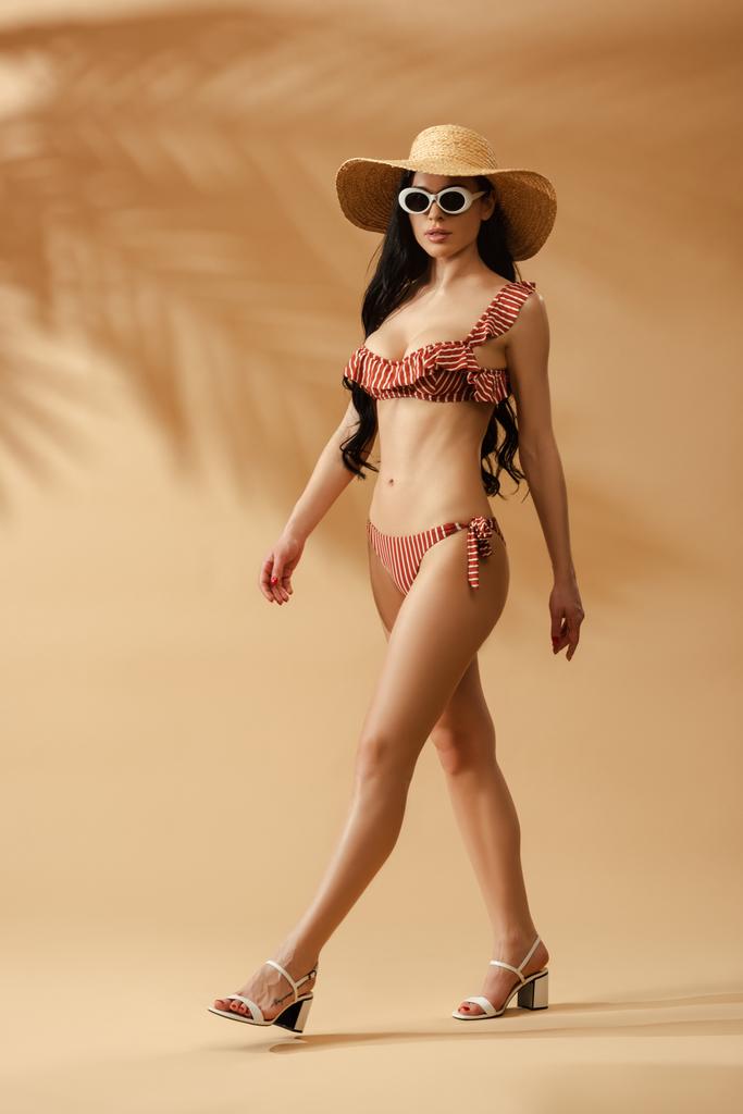 Çizgili mayo, hasır şapka ve güneş gözlüğü takan, bej arka planda yürüyen seksi esmer kadın. - Fotoğraf, Görsel
