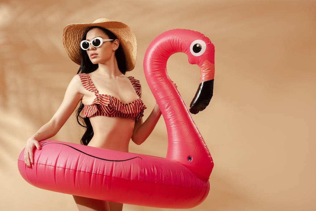 sexy brunette vrouw in gestreepte badpak, zonnebril en stro hoed met opblaasbare flamingo op beige achtergrond - Foto, afbeelding