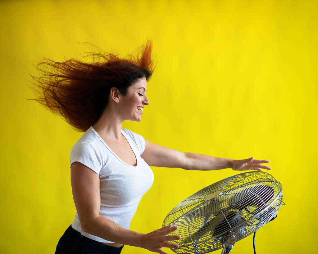 Güzel kızıl saçlı bir kadın, sarı arka planda büyük bir elektrikli vantilatörün başında serinliyor. Saçları rüzgarda savrulan bir kız. Havayı soğutmak için aygıt. - Fotoğraf, Görsel
