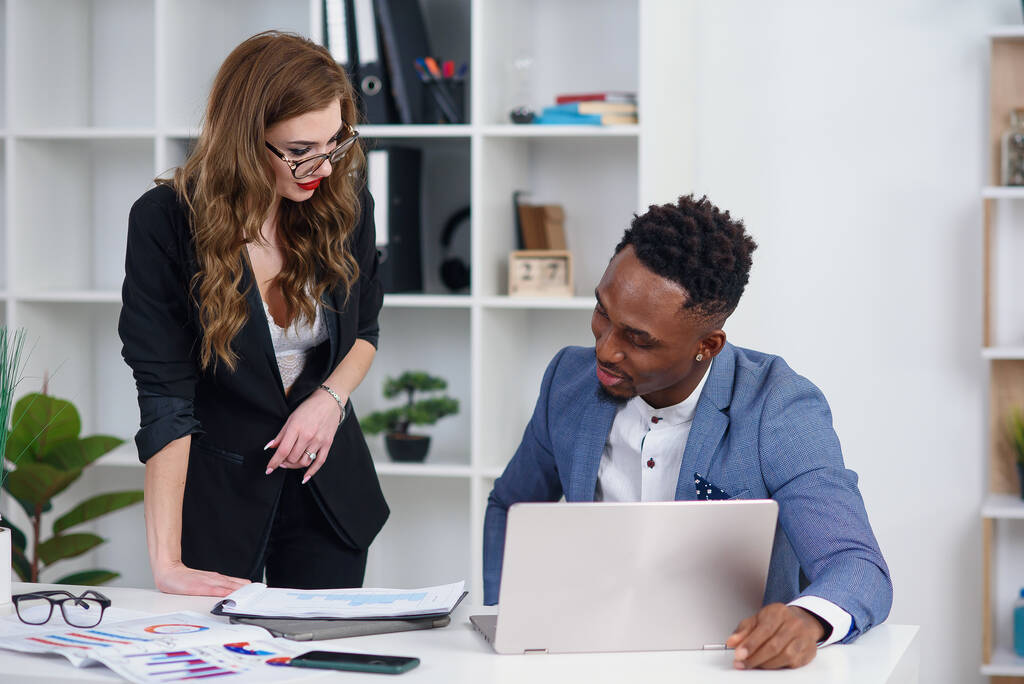 Two diverse businesspeople chatting sitting behind laptop in office. Excited caucasian female sharing ideas or startup business plan with black male coworker. Informal conversation, work break concept - Photo, Image