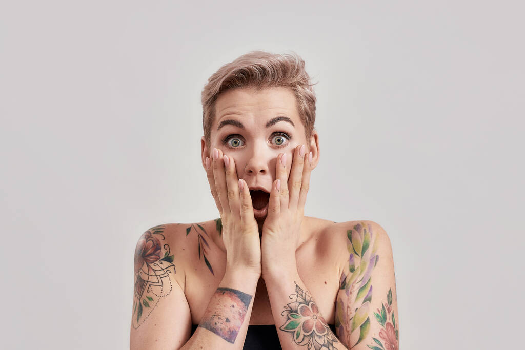Hämmästyttävää Muotokuva hämmästynyt tatuoitu nainen lyhyet hiukset näyttävät hämmästynyt tai järkyttynyt kameran suu auki, koskettaa kasvot eristetty valon taustalla - Valokuva, kuva