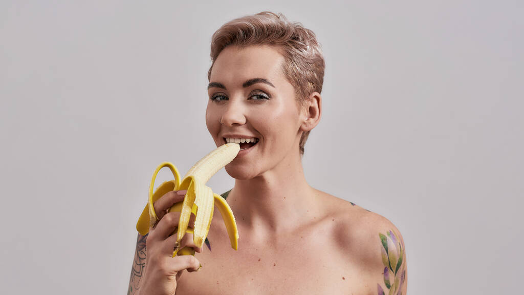 Houkutteleva tatuoitu nainen, jolla on lävistetty nenä ja lyhyet hiukset, katselee kameraa, maistellen, syöden banaania eristettynä harmaalla taustalla - Valokuva, kuva