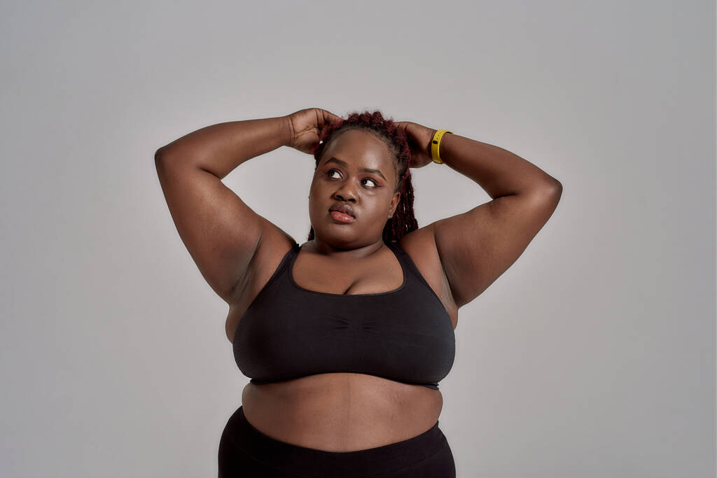 Şişkin, ayrıca siyah spor giyim giyen Afrikalı Amerikalı kadın stüdyoda gri arka planda dikilirken düşünceli bir şekilde yana bakıyor. Spor anlayışı, sağlıklı yaşam tarzı, vücut pozitifliği, eşitlik - Fotoğraf, Görsel