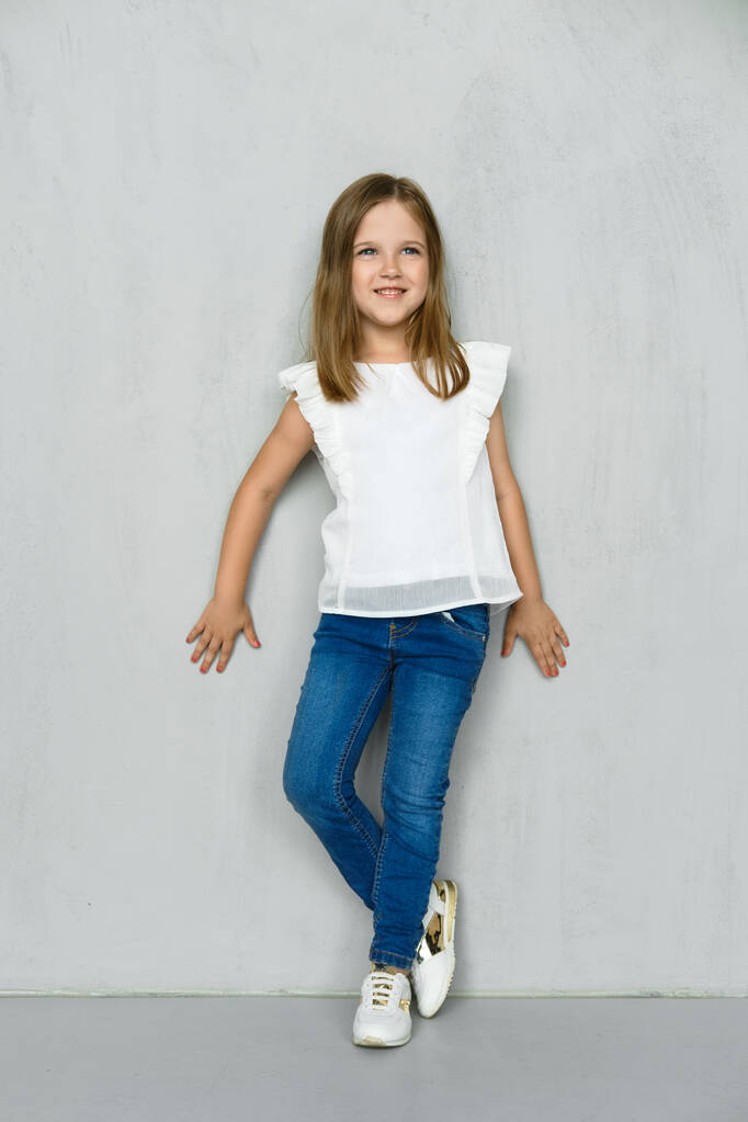 Маленька дівчинка в білій блузці і джинсах, що схиляються до стіни в студії
 - Фото, зображення
