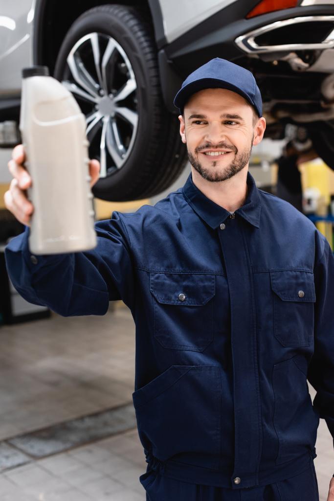 selective focus of happy mechanic in cap and uniform holding bottle with car oil  - Photo, Image