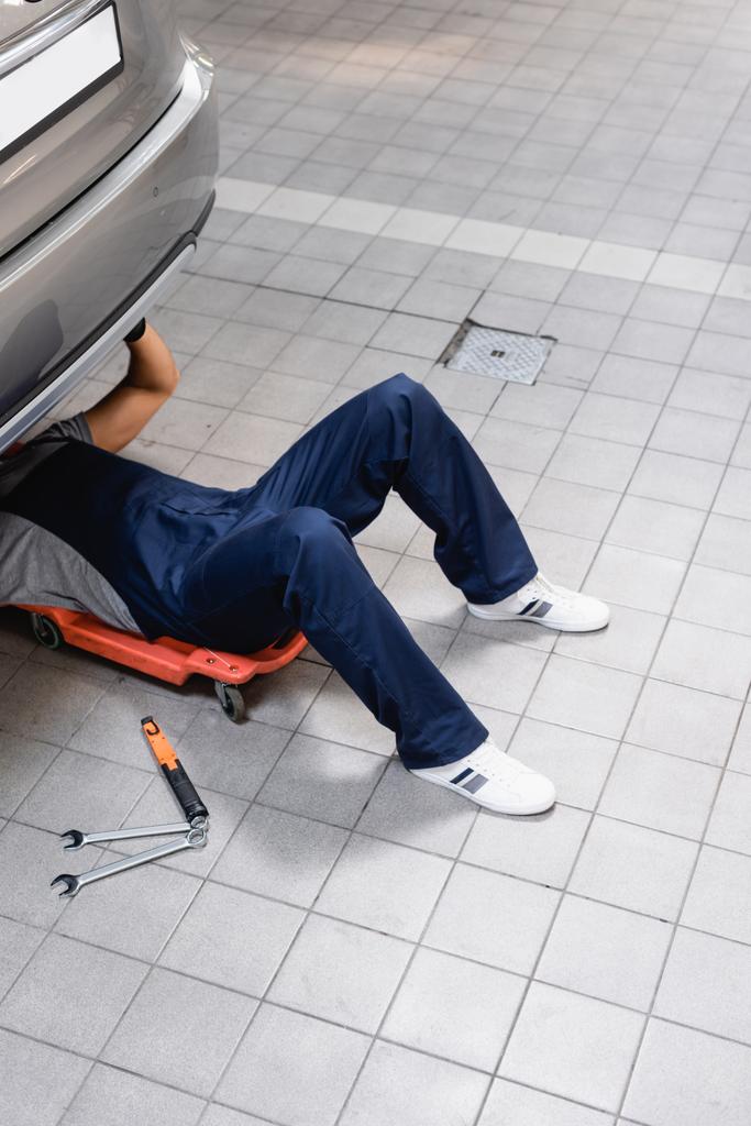 mecânico em uniforme deitado sob carro perto de ferramentas metálicas
 - Foto, Imagem