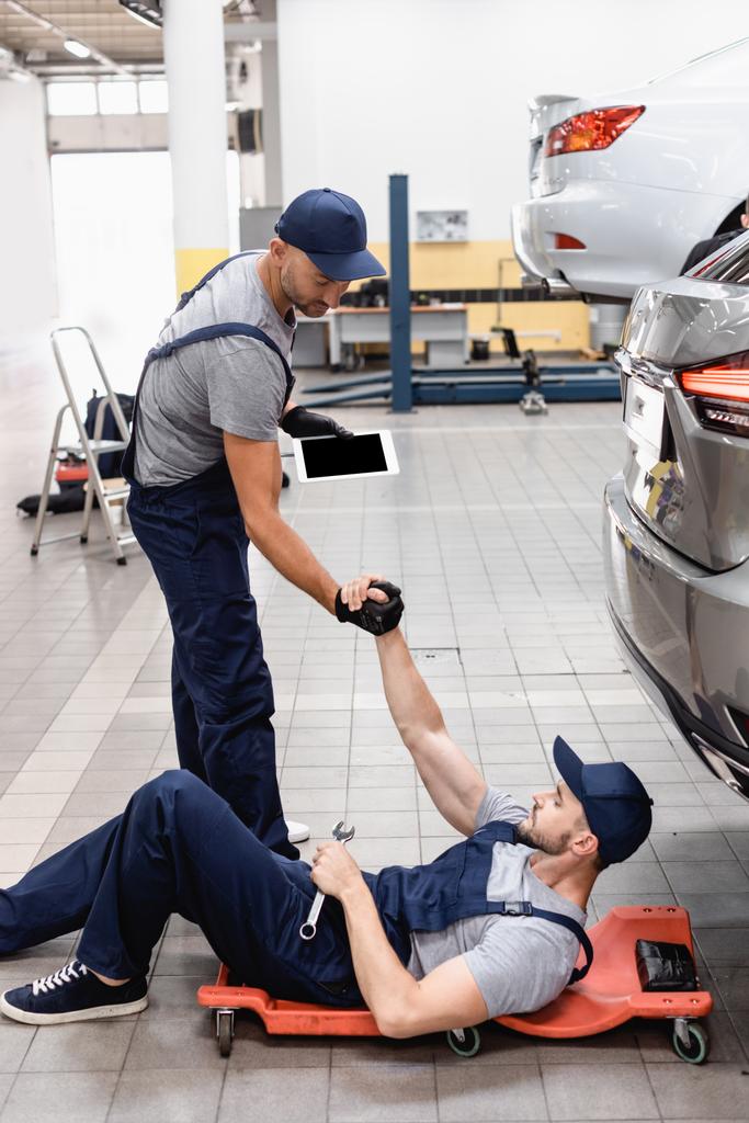 mechanic with digital tablet holding hands with coworker in cap lying near car - Photo, Image