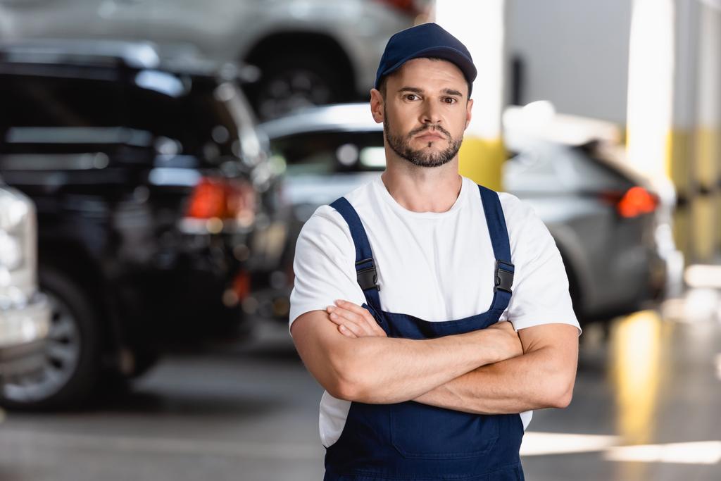 handsome mechanic in uniform and cap standing with crossed arms in car service - Photo, Image