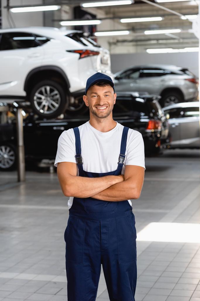 onnellinen mekaanikko univormussa ja korkki seisoo ristissä kädet huoltoasemalla - Valokuva, kuva