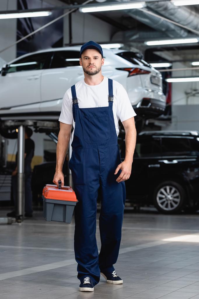 handsome mechanic in overalls and cap holding toolbox in service station - Photo, Image
