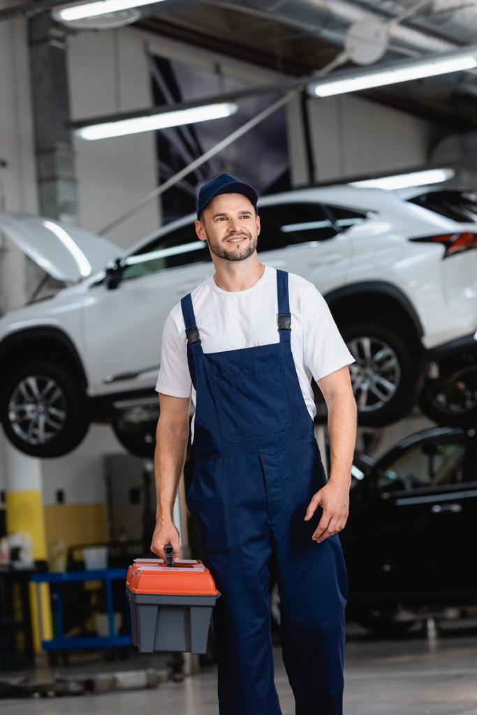 gelukkige monteur in overalls en cap holding toolbox in de auto service  - Foto, afbeelding