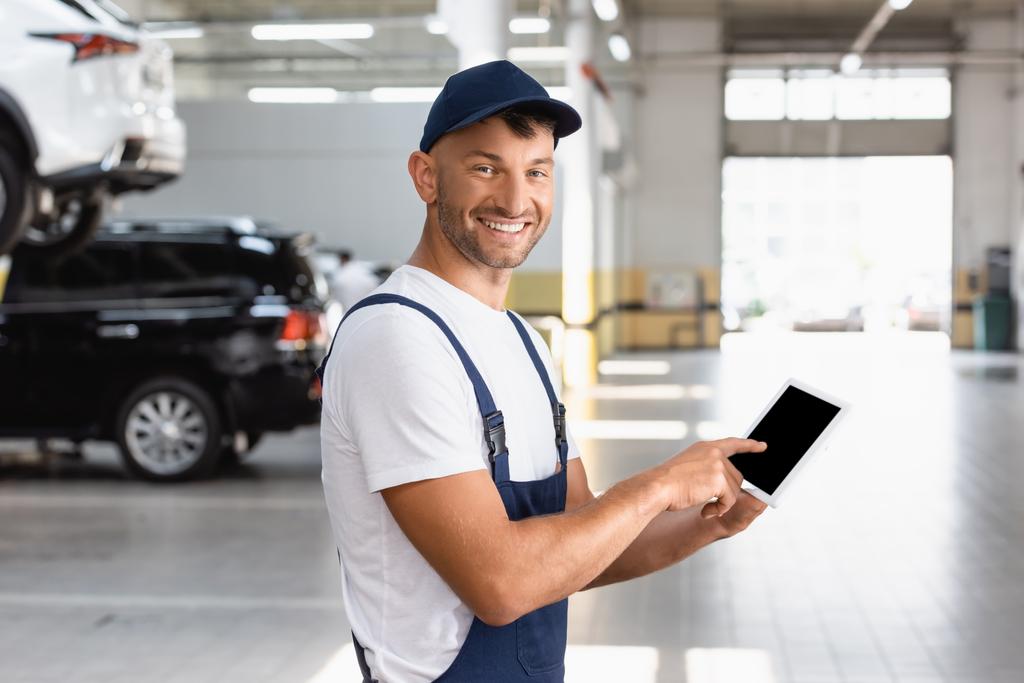 glücklicher Mechaniker in Overalls und Mütze zeigt mit dem Finger auf digitales Tablet mit leerem Bildschirm im Autoservice  - Foto, Bild