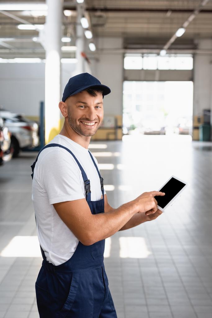 cheerful mechanic in overalls and cap pointing with finger at digital tablet with blank screen in car service  - Photo, Image