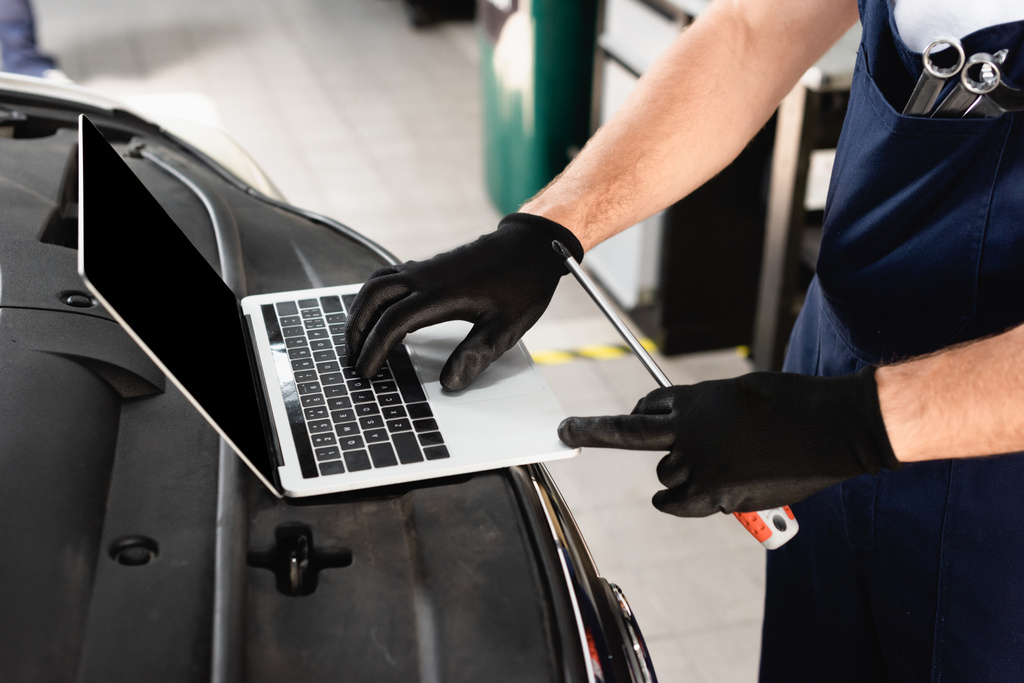 Gesneden weergave van auto monteur typen op laptop op auto en met schroevendraaier op tankstation - Foto, afbeelding