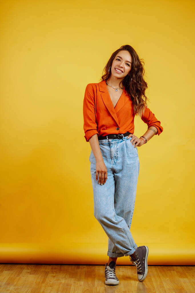 Portret van een jong meisje dat poseert op een gele achtergrond. Mooie vrouw draagt een wortel blouse en jeans. Ontwerp van mode en levensstijl. - Foto, afbeelding