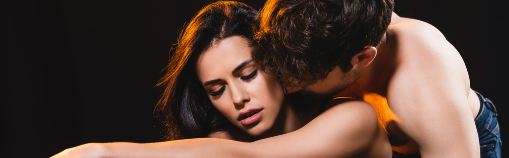 panoramic crop of muscular man kissing woman with closed eyes isolated on black  - Photo, Image
