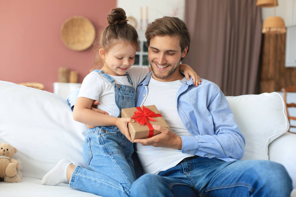 Χαριτωμένη μικρή κόρη κάνει έκπληξη στον μπαμπά, κοριτσάκι παρουσιάζει κουτί δώρου στον πατέρα κάθονται στον καναπέ. Ημέρα του πατέρα - Φωτογραφία, εικόνα