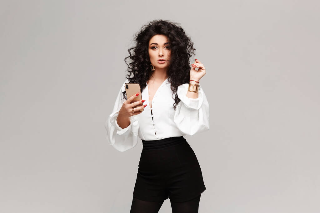 Retrato de moda de uma menina modelo em blusa branca e shorts pretos com um smartphone na mão olhando para a câmera no fundo branco, isolado
 - Foto, Imagem