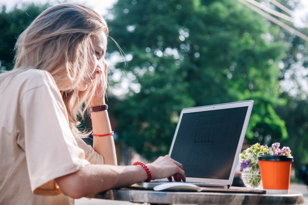 Een jong meisje op een zomerspeelplaats van een coffeeshop werkt achter een laptop en drinkt koffie uit een wegwerpbeker  - Foto, afbeelding