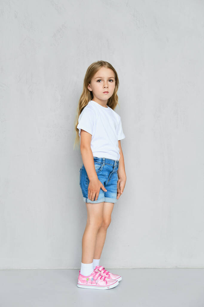 Malá holčička v bílém tričku, džínách a růžových teniskách, stojící v profilu u zdi - Fotografie, Obrázek