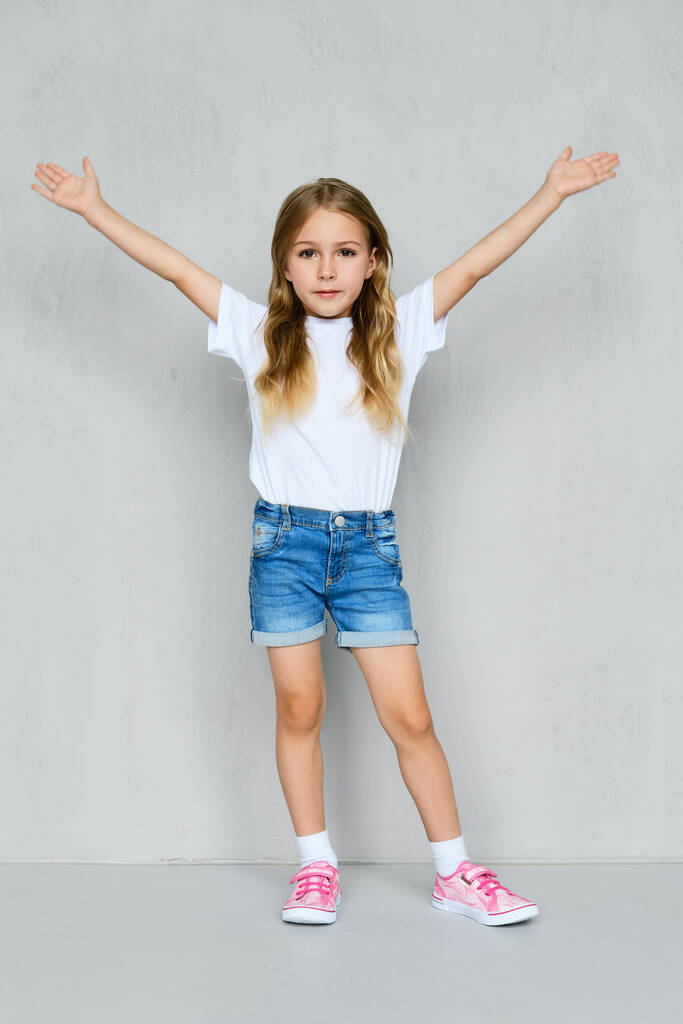 Klein meisje in wit t-shirt, jeans short en roze sneakers met opgeheven handen bij de muur - Foto, afbeelding