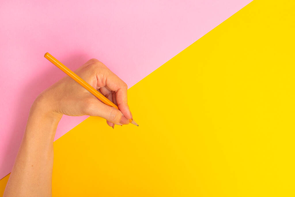 La mano sinistra tiene una matita e sta per scrivere qualcosa su uno sfondo rosa-giallo brillante. Giornata internazionale dei mancini il 13 agosto - Foto, immagini