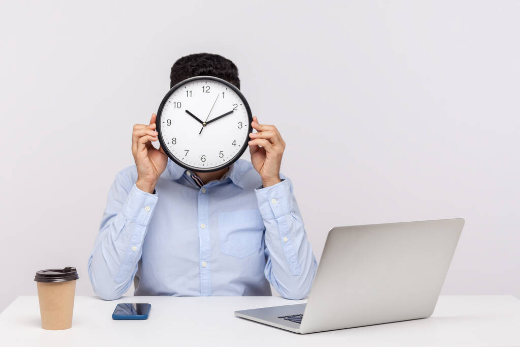 Zeit zu arbeiten! Männliche Mitarbeiter sitzen am Büroarbeitsplatz und bedecken das Gesicht mit großer Uhr, Besprechungstermin, Zeitmanagement-Konzept, Geschäftsplan. Innenstudio isoliert auf weißem Hintergrund aufgenommen - Foto, Bild