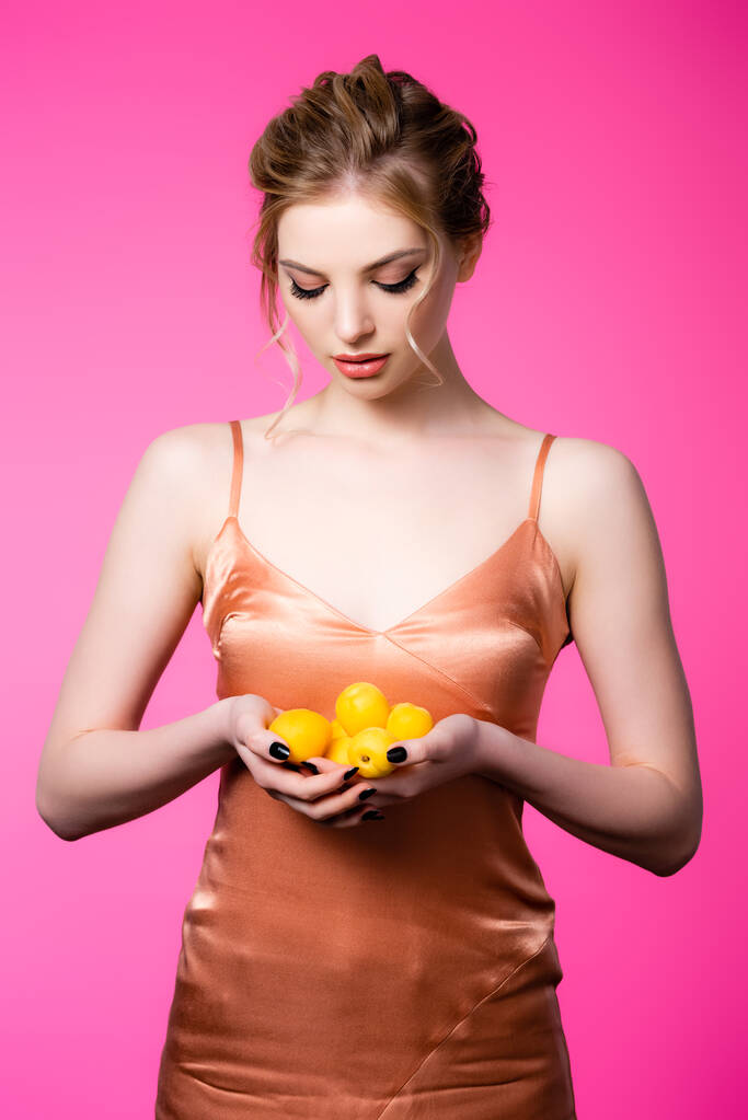 elegante schöne blonde Frau im Seidenkleid mit Blick auf reife Aprikosen isoliert auf rosa - Foto, Bild