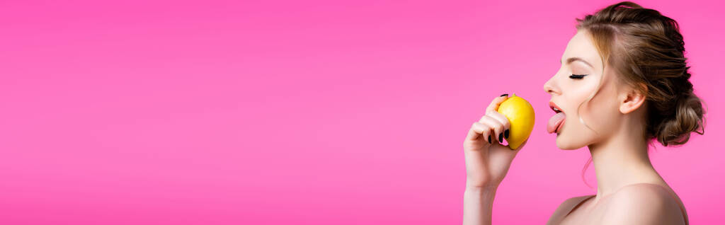 zijaanzicht van mooie blonde vrouw likken rijp citroen geïsoleerd op roze, panoramisch schot - Foto, afbeelding
