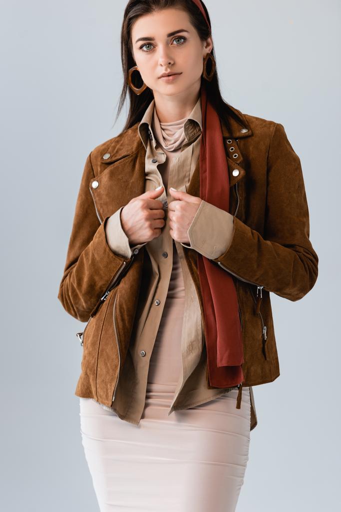confident, stylish girl touching suede jacket while looking at camera isolated on grey - Photo, Image