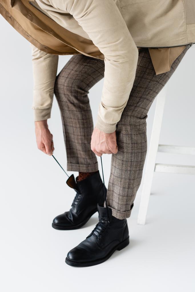 abgeschnittene Ansicht eines modischen Mannes, der Schnürsenkel an schwarzen Stiefeln bindet, während er auf einem Schemel auf weißem sitzt - Foto, Bild