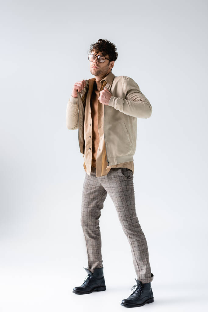 full length view of stylish man touching jacket while looking at camera on grey - Photo, Image
