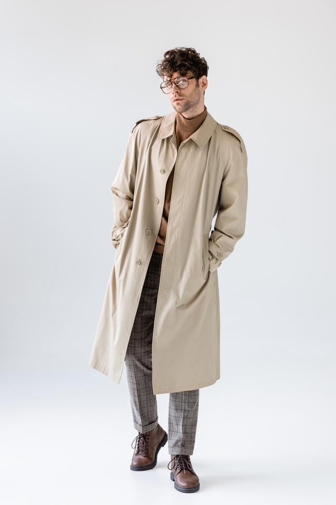 Вид в полный рост модного мужчины в трикотажном пальто, держащего руки в карманах и позирующего на сером - Фото, изображение
