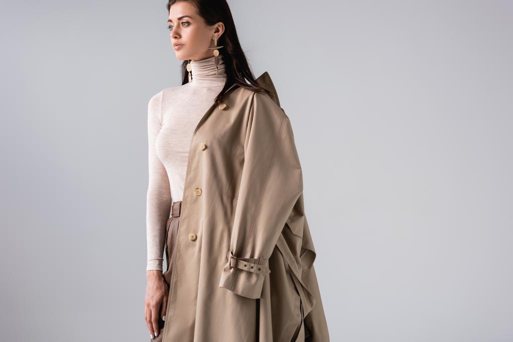 attractive, trendy woman with trench coat on shoulder posing isolated on grey - Photo, Image