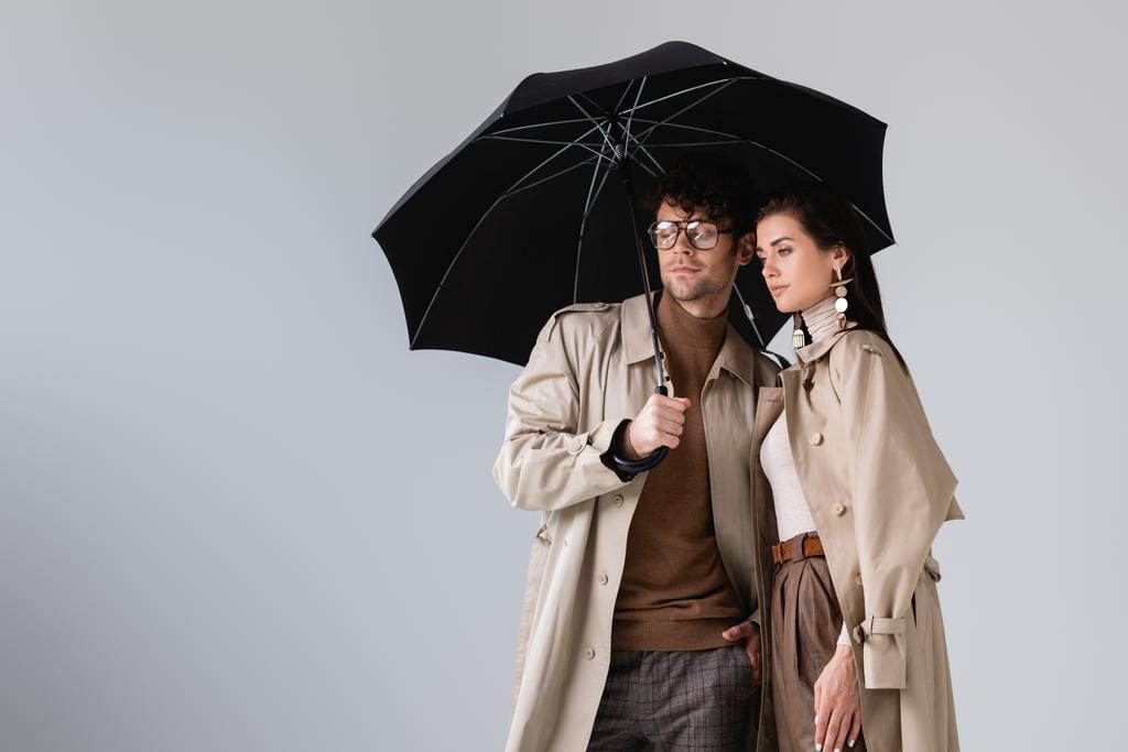 fiatal, divatos pár őszi ruhában pózol esernyő alatt, miközben félrenéz elszigetelve szürke - Fotó, kép