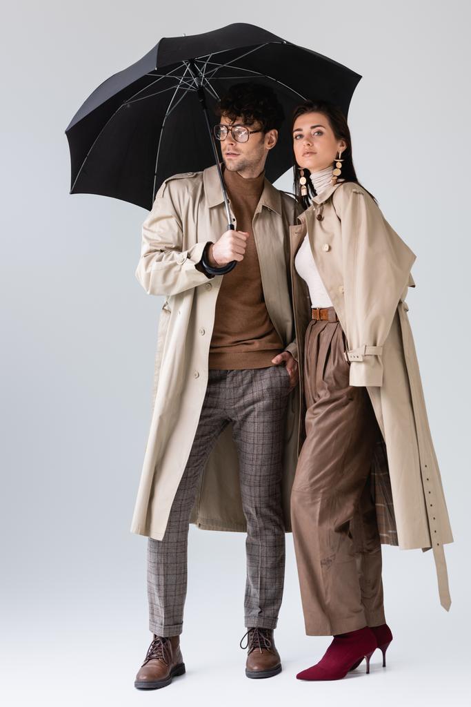 volledige weergave van jong paar in modieuze herfst kleding poseren onder paraplu op grijs - Foto, afbeelding