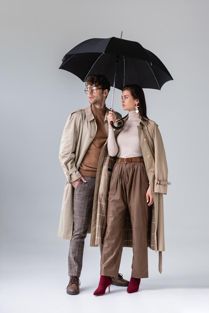 vista completa de la pareja con estilo en ropa de otoño de pie bajo el paraguas mientras mira hacia otro lado en gris
 - Foto, Imagen