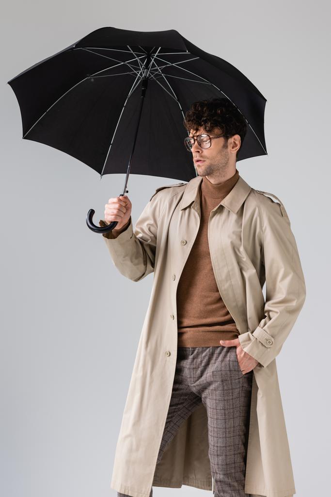 стильный мужчина, стоящий под черным зонтиком с закрытыми глазами, изолированными на сером - Фото, изображение
