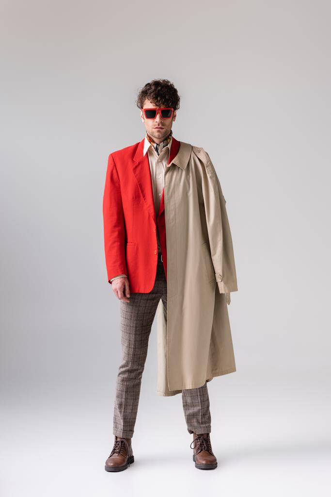 visão de comprimento total da moda, homem confiante com casaco de trincheira sobre ombro olhando para a câmera no cinza
 - Foto, Imagem