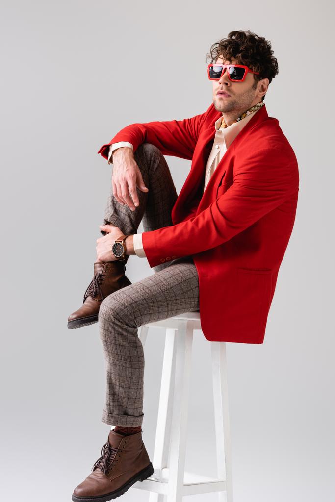 μοντέρνα άνθρωπος σε κόκκινο σακάκι και γυαλιά ηλίου αγγίζοντας το πόδι, ενώ κάθεται σε σκαμνί απομονώνονται σε γκρι - Φωτογραφία, εικόνα