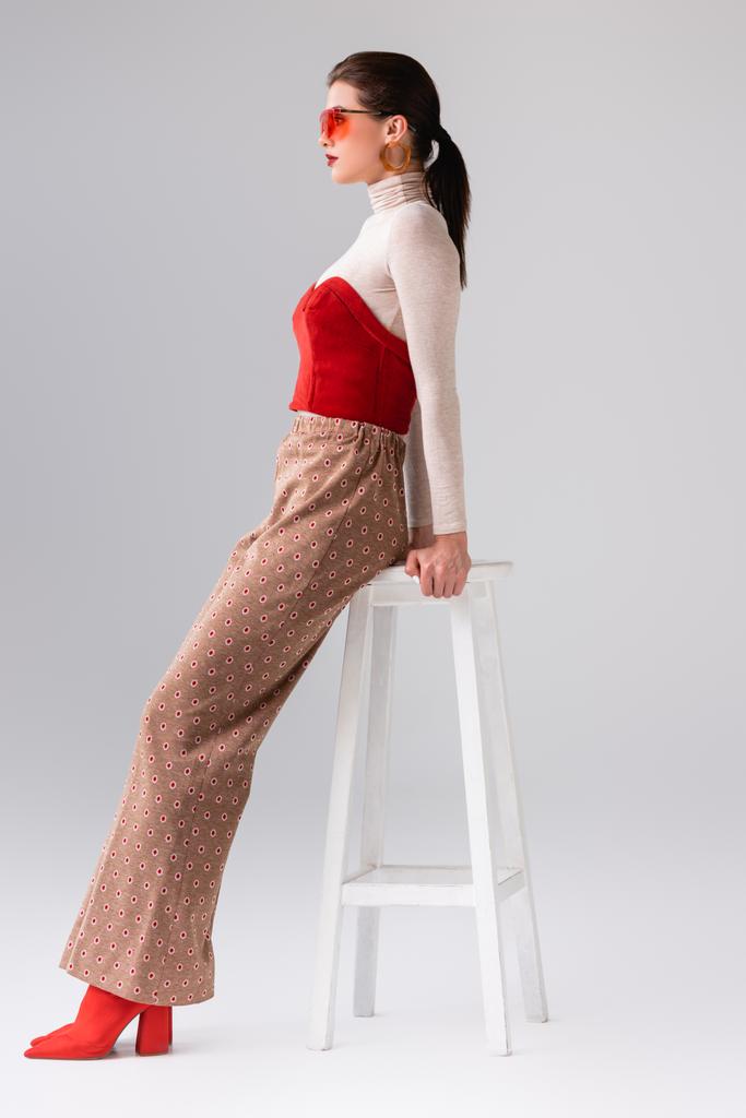 side view of fashionable girl in beige trousers and red corset on turtleneck leaning on stool on grey - Photo, Image