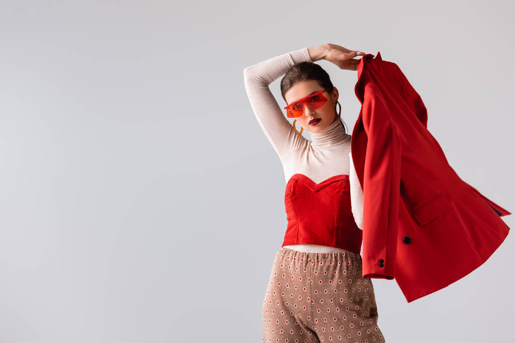 divatos lány napszemüvegben kezében piros blézer, miközben néz kamera elszigetelt szürke - Fotó, kép