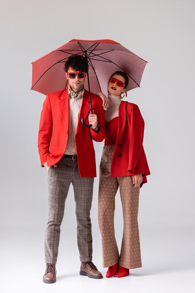vue pleine longueur de jeune couple branché en blazers rouges posant avec parapluie sur gris - Photo, image