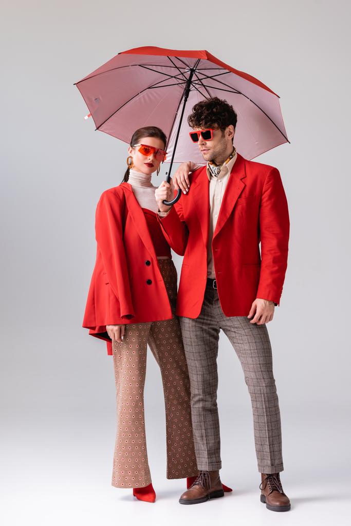 Ganzkörperansicht des trendigen Paares in roten Blazern und Sonnenbrille posiert mit Sonnenschirm auf grau - Foto, Bild