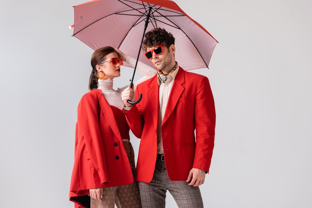 mladý, módní pár v červených blejzrech a slunečních brýlích pózující pod deštníkem izolovaným na šedi - Fotografie, Obrázek